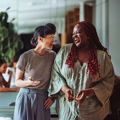 Two women laughing together in an office setting, one in grey shirt and one in mint green cardigan with red beaded necklace.