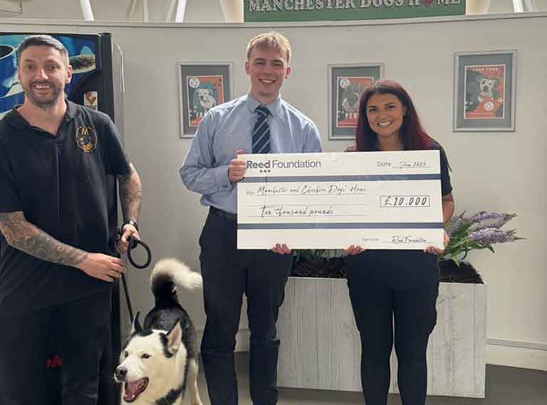 Three people pose with a large ceremonial cheque for ten thousand pounds, with a husky dog on the left side of the group.