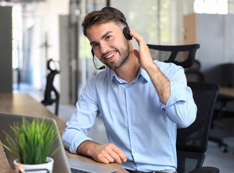 Smiling male call-centre operator with headphones sitting at modern office, consulting online information in a laptop, looking up information in a file in order to be of assistance to the client.