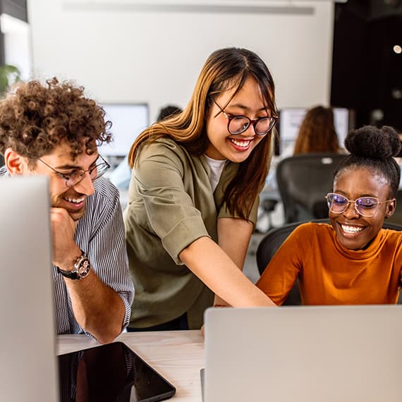 Three diverse coworkers smiling while collaborating at a laptop in a bright modern office setting.