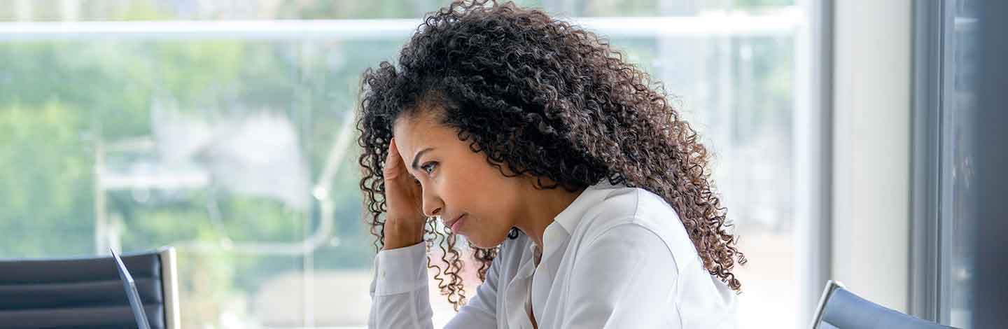 Woman with curly hair in white shirt looking stressed with hand on forehead in bright office setting.