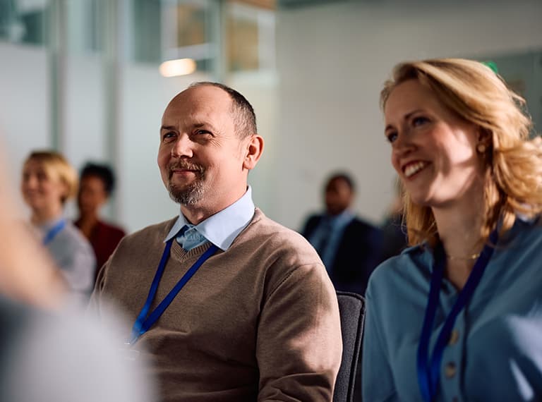 Two professionals with blue lanyards smiling while attending a business presentation or conference in a modern office.