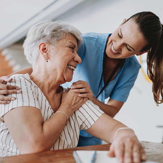 Young caregiver in blue scrubs smiling with senior woman with short white hair wearing striped shirt in bright room