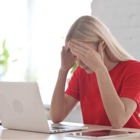 Woman in red shirt covering her face with hands while sitting at laptop, appearing stressed or upset.