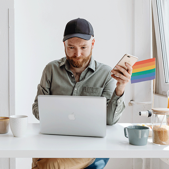 Bearded man in black cap and gray shirt looking at phone while sitting at white desk with laptop and coffee mug.