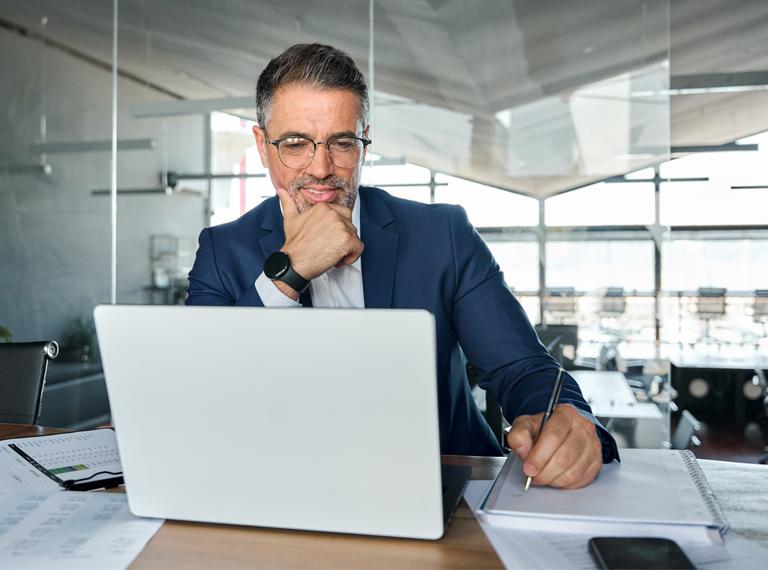 Middle aged business man executive wearing suit sitting at desk working on laptop writing notes. Professional businessman ceo manager using computer for elearning watching webinar in modern office.