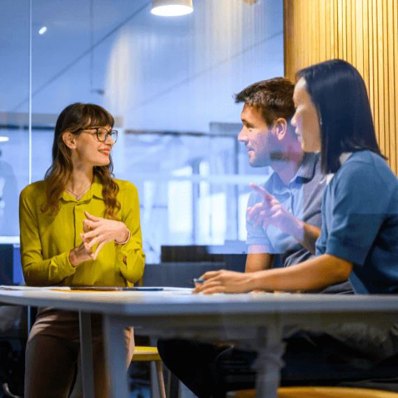Three professionals having a discussion at a table in a modern office with blue and yellow lighting accents.