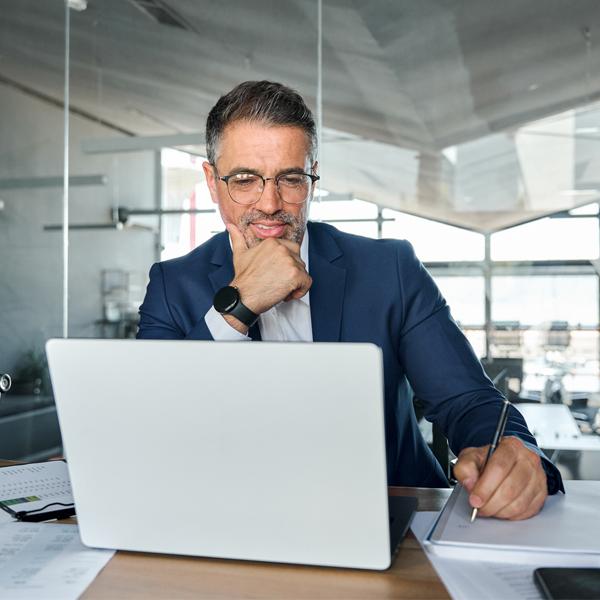 Middle aged business man executive wearing suit sitting at desk working on laptop writing notes. Professional businessman ceo manager using computer for elearning watching webinar in modern office.