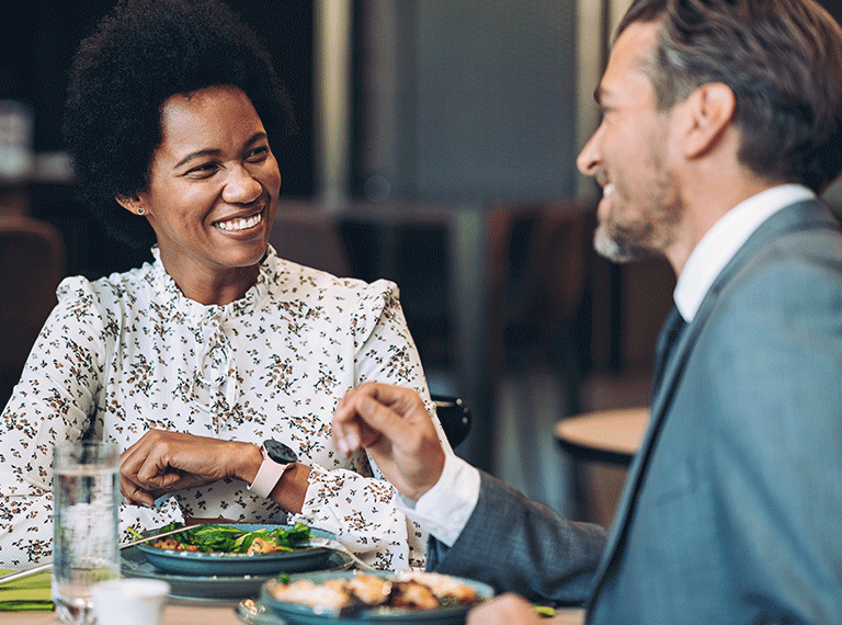 Woman with natural hair in floral blouse smiling at man in gray suit during business lunch meeting with salads on table.