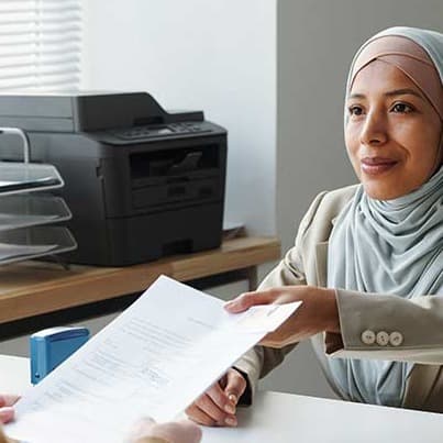 Woman in hijab hands document to person across desk in bright office with printer and file organizer in background.
