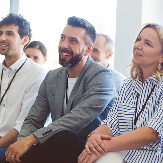 Three smiling professionals sitting in a bright room during a presentation, with a bearded man in gray blazer at center.
