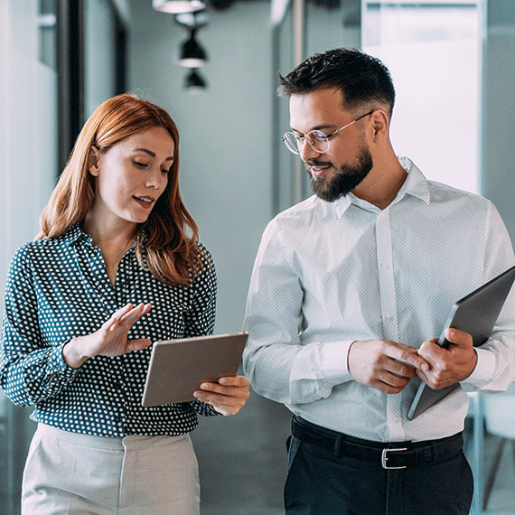 Two business professionals reviewing documents together in a modern office hallway, woman in polka dot blouse and man in glasses.