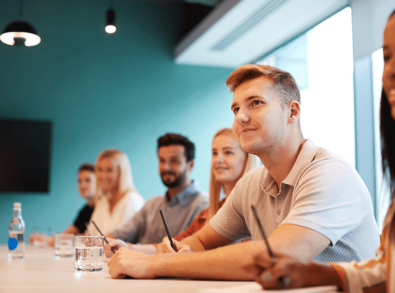 Young man in beige shirt smiling while sitting at a conference table with colleagues in a modern office setting.