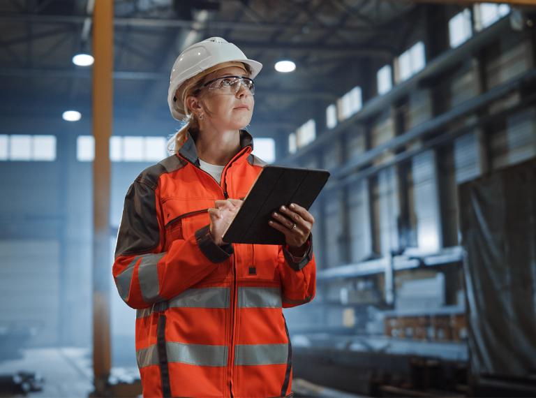 Professional Heavy Industry Engineer Worker Wearing Safety Uniform and Hard Hat, Using Tablet Computer. Serious Successful Female Industrial Specialist Walking in a Metal Manufacture Warehouse.