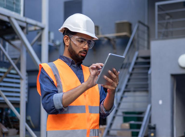 Serious thinking and focused engineer working in factory in hard hat and vest, man using tablet computer reading diagram, and checking equipment, inside industrial factory.