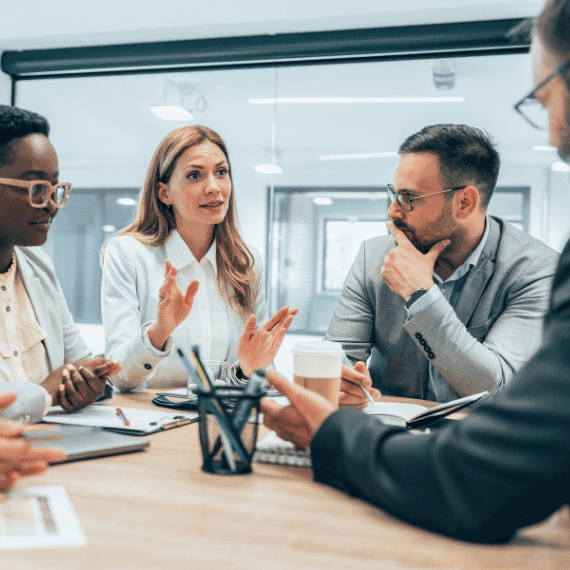 Diverse business team in meeting, woman in white shirt gesturing while speaking to attentive colleagues around table.