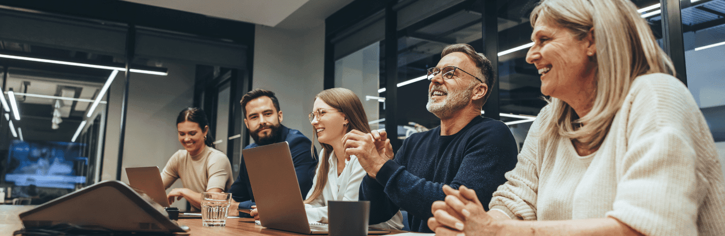 Five diverse professionals smiling and collaborating around a conference table with laptops in a modern office setting.