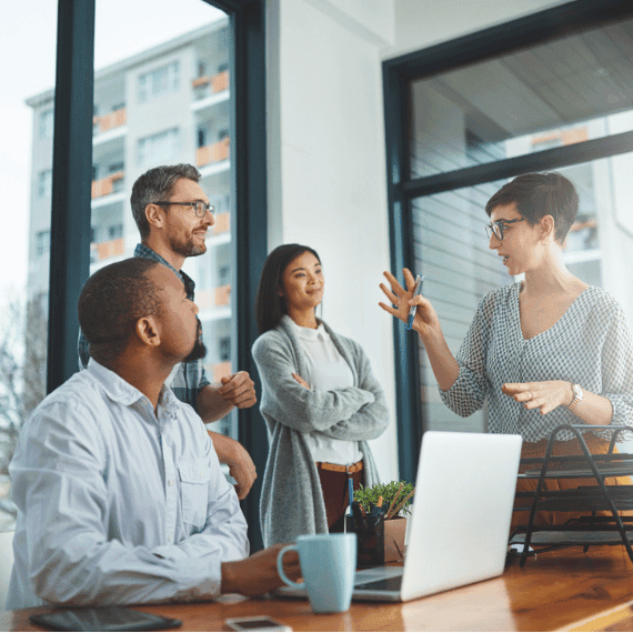 Four diverse professionals collaborating in a modern office with large windows, laptop on wooden table, engaged in discussion.