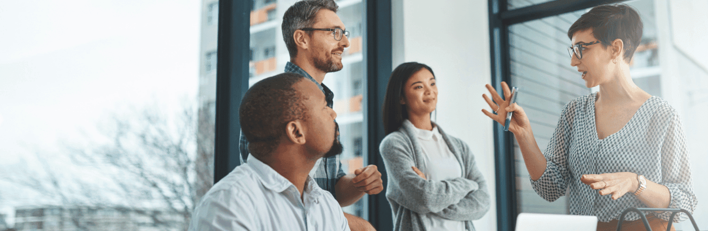 Four diverse professionals engaged in discussion in modern office with large windows and natural light.