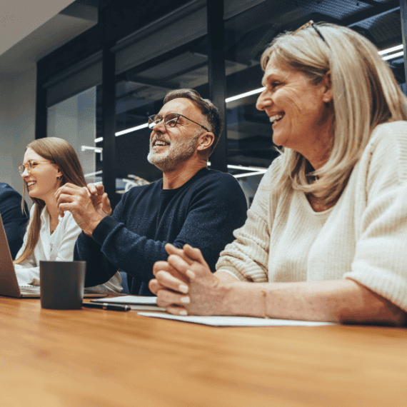 Three smiling professionals sitting at a wooden table in a modern office, engaged in a meeting or presentation.