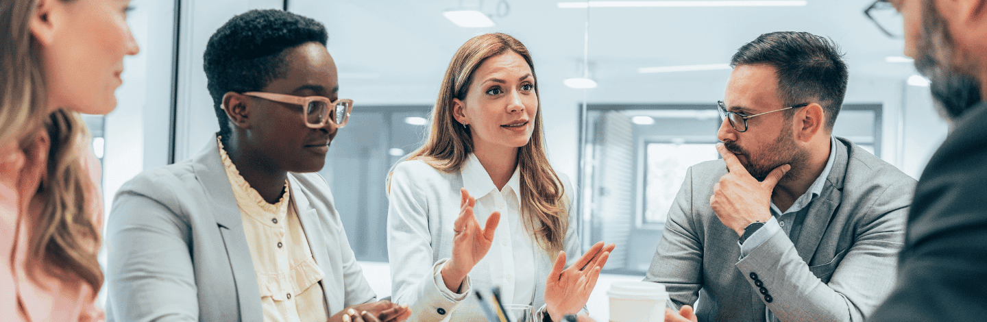 Woman in white blazer speaking with hand gestures during business meeting with diverse colleagues in modern office.